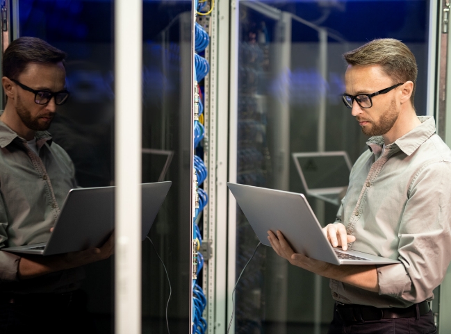 A Caucasian man with brown hair and glasses studies his laptop screen while inspecting a data center for safety measures.